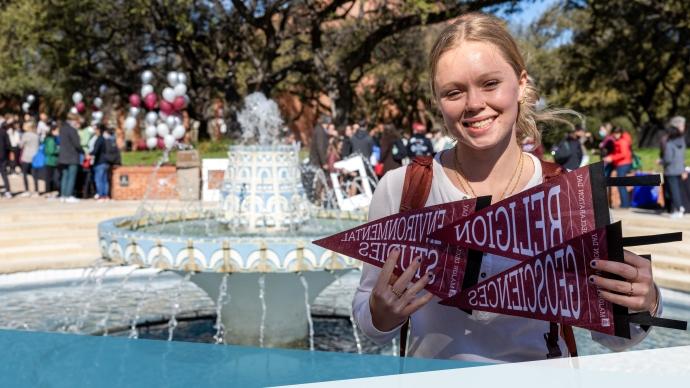 Student holding up degree pennants for Religion, Geosciences, and Environmental Studies in front of Miller fountain