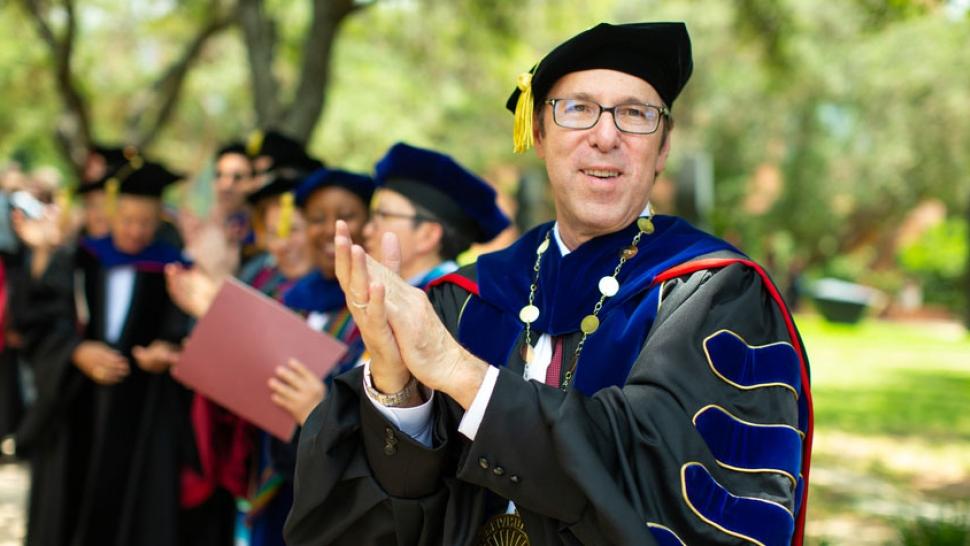 Danny Anderson, in graduation robes, claps at graduation, next to professors in graduation robes. 