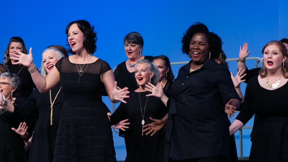 rosalind jones does jazz hands in her all-female a cappella group. all members are on stage wearing black against a blue backdrop